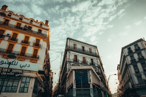 The scene depicts several historic buildings with intricate balconies lining a narrow street. The architecture features vibrant colors with orange-tinted details, and the sky overhead is partially cloudy with a warm glow. Arabic signage is visible, suggesting a Middle Eastern or North African urban setting.