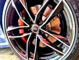 A close-up of a car wheel featuring a modern, sporty alloy rim design with multiple sharp angles and glossy finish. The brake caliper behind the rim is painted in a vivid red color, and the details of the brake disc are visible with perforations. The logo of a well-known automobile brand is prominently displayed at the center of the rim.