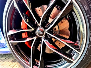 A close-up of a car wheel featuring a modern, sporty alloy rim design with multiple sharp angles and glossy finish. The brake caliper behind the rim is painted in a vivid red color, and the details of the brake disc are visible with perforations. The logo of a well-known automobile brand is prominently displayed at the center of the rim.