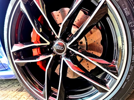 A close-up of a car wheel featuring a modern, sporty alloy rim design with multiple sharp angles and glossy finish. The brake caliper behind the rim is painted in a vivid red color, and the details of the brake disc are visible with perforations. The logo of a well-known automobile brand is prominently displayed at the center of the rim.