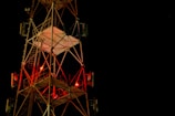 A tall metal communications tower is illuminated against a dark night sky. Red lights are visible on the structure, and it is composed of a complex network of beams and ladders. The tower's geometric design stands out sharply against the deep black background.