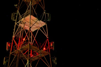 A tall metal communications tower is illuminated against a dark night sky. Red lights are visible on the structure, and it is composed of a complex network of beams and ladders. The tower's geometric design stands out sharply against the deep black background.