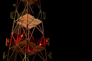 A tall metal communications tower is illuminated against a dark night sky. Red lights are visible on the structure, and it is composed of a complex network of beams and ladders. The tower's geometric design stands out sharply against the deep black background.
