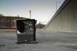 A broken television set is placed on a gravel area near a large concrete wall. The TV screen is shattered, and surrounding debris can be seen. In the background, there are residential buildings and a few trees, along with streetlights and electrical poles. The lighting suggests it’s either early morning or late afternoon.