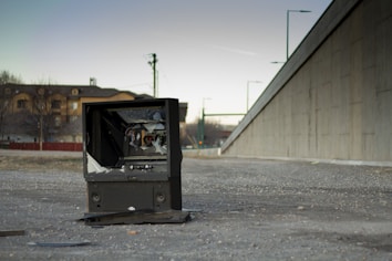 A broken television set is placed on a gravel area near a large concrete wall. The TV screen is shattered, and surrounding debris can be seen. In the background, there are residential buildings and a few trees, along with streetlights and electrical poles. The lighting suggests it’s either early morning or late afternoon.