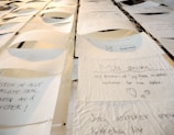 A collection of handwritten notes on thin, textured paper, suspended and displayed in rows. Each note contains text and small drawings, with varying handwriting styles. The setting appears artistic, with a focus on personal expressions or wishes.