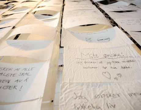 A collection of handwritten notes on thin, textured paper, suspended and displayed in rows. Each note contains text and small drawings, with varying handwriting styles. The setting appears artistic, with a focus on personal expressions or wishes.