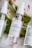Three bottles of face mist labeled with different ingredients, placed on a surface with pink and white flowers in the background. The central bottle is labeled as Aloe + Rosewater Hydrating Face Mist.