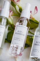Three bottles of face mist labeled with different ingredients, placed on a surface with pink and white flowers in the background. The central bottle is labeled as Aloe + Rosewater Hydrating Face Mist.