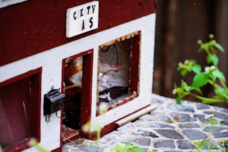 A miniature structure resembling a house with a sign that reads 'Creativ Haus'. The walls are painted red and white, and there are windows with wire details inside. The scene includes cobblestone paving and some green plants with small flowers in the foreground.