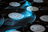 Multiple vinyl records are scattered across a surface, each showing distinctive labels and grooves. The lighting casts a bluish tint, highlighting the shiny, reflective surface of the records.