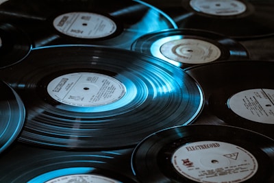 Multiple vinyl records are scattered across a surface, each showing distinctive labels and grooves. The lighting casts a bluish tint, highlighting the shiny, reflective surface of the records.