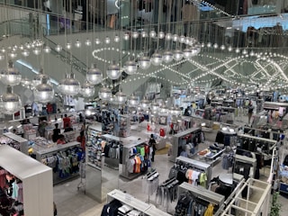 A busy clothing store interior with multiple sections displaying various types of clothing. Shiny spherical light fixtures hang from the ceiling in a mirrored setting, creating a reflective effect throughout the space. Customers are browsing through racks of clothes, which include shirts, pants, and other garments.