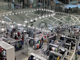 A busy clothing store interior with multiple sections displaying various types of clothing. Shiny spherical light fixtures hang from the ceiling in a mirrored setting, creating a reflective effect throughout the space. Customers are browsing through racks of clothes, which include shirts, pants, and other garments.