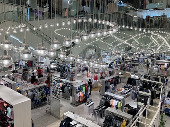 A busy clothing store interior with multiple sections displaying various types of clothing. Shiny spherical light fixtures hang from the ceiling in a mirrored setting, creating a reflective effect throughout the space. Customers are browsing through racks of clothes, which include shirts, pants, and other garments.