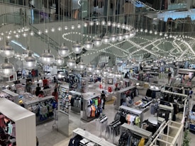 A busy clothing store interior with multiple sections displaying various types of clothing. Shiny spherical light fixtures hang from the ceiling in a mirrored setting, creating a reflective effect throughout the space. Customers are browsing through racks of clothes, which include shirts, pants, and other garments.