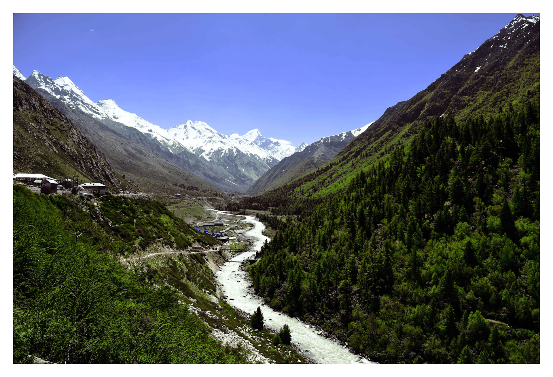 A serene view of the crystal-clear river flowing through the lush green Sangla Valley, with traditional wooden houses dotting the hillside.