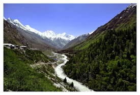 A serene valley with a meandering river flowing through lush green hills. Snow-capped mountains stand majestically in the background under a clear blue sky. Some houses are visible along the hills, adding a touch of habitation to the natural landscape.