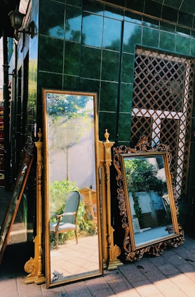 Two ornate mirrors with golden frames are propped up against a dark green tiled wall. The reflections in the mirrors show a blue chair and some greenery, indicating the presence of a garden or outdoor space. The setting appears to be outdoors, possibly in a street market or antique shop area.