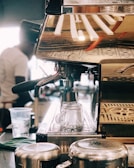 A close-up view of a coffee machine with a metallic finish, reflecting the environment around it. Two glass cups are positioned in the drip area, while a cloth and metal jugs are nearby. A blurred figure in the background appears to be operating the machine, suggesting a bustling coffee shop environment.