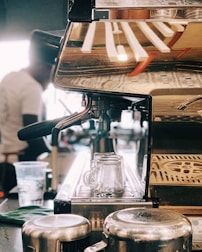 A close-up view of a coffee machine with a metallic finish, reflecting the environment around it. Two glass cups are positioned in the drip area, while a cloth and metal jugs are nearby. A blurred figure in the background appears to be operating the machine, suggesting a bustling coffee shop environment.