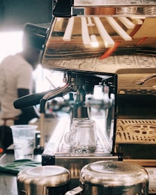 A close-up view of a coffee machine with a metallic finish, reflecting the environment around it. Two glass cups are positioned in the drip area, while a cloth and metal jugs are nearby. A blurred figure in the background appears to be operating the machine, suggesting a bustling coffee shop environment.