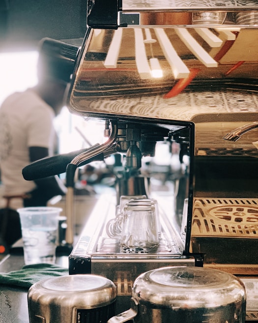 A close-up view of a coffee machine with a metallic finish, reflecting the environment around it. Two glass cups are positioned in the drip area, while a cloth and metal jugs are nearby. A blurred figure in the background appears to be operating the machine, suggesting a bustling coffee shop environment.