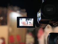 A video camera captures a scene with seats and a person in white attire visible through the camera's display screen. The surroundings are blurred and have a warm color tone.