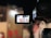 A video camera captures a scene with seats and a person in white attire visible through the camera's display screen. The surroundings are blurred and have a warm color tone.
