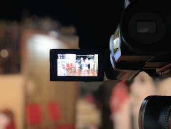 A video camera captures a scene with seats and a person in white attire visible through the camera's display screen. The surroundings are blurred and have a warm color tone.