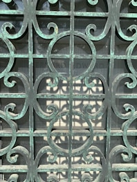 An ornate metal gate with a patina finish, featuring intricate scrollwork patterns. Behind the gate, there is a stone slab with partially readable engraving.