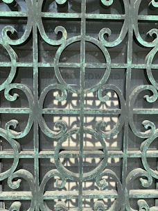 An ornate metal gate with a patina finish, featuring intricate scrollwork patterns. Behind the gate, there is a stone slab with partially readable engraving.