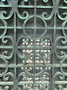 An ornate metal gate with a patina finish, featuring intricate scrollwork patterns. Behind the gate, there is a stone slab with partially readable engraving.