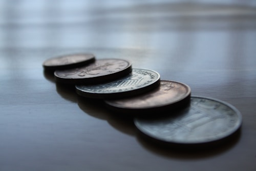 Several coins are placed on a reflective surface, aligned in a staggered manner. The coins cast soft shadows on the surface, and the image has a shallow depth of field, focusing on the coins while the background is blurred.