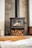A modern wood-burning stove with a glass front, set against a contemporary tiled wall. Below the stove, a stack of neatly arranged logs is visible. To the side, decorative vases and a pot add to the aesthetic. The warm tones of the wood and the sleek design create an inviting atmosphere.