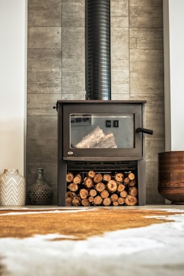 A modern wood-burning stove with a glass front, set against a contemporary tiled wall. Below the stove, a stack of neatly arranged logs is visible. To the side, decorative vases and a pot add to the aesthetic. The warm tones of the wood and the sleek design create an inviting atmosphere.