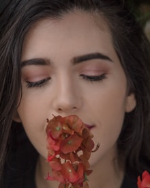 A person with long dark hair and closed eyes holds an orange-red flower close to their face. The petals of the flower are prominently visible, and the person appears calm and serene.