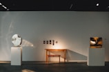An art gallery space with a minimalistic design featuring two sculptures on white pedestals. The left sculpture is circular with a reflective surface, while the right sculpture has an abstract, angular form with a golden hue. A simple wooden bench is placed against the wall between the sculptures, accompanied by small framed artworks hung above.