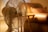A vintage-style table fan with a metal grill is in focus, placed in a warmly lit room. Behind the fan, an out-of-focus, cushioned chair and a side table with a soft, ambient light from possibly a lamp create a cozy atmosphere.