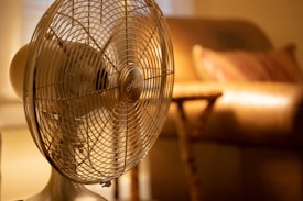 A vintage-style table fan with a metal grill is in focus, placed in a warmly lit room. Behind the fan, an out-of-focus, cushioned chair and a side table with a soft, ambient light from possibly a lamp create a cozy atmosphere.