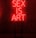 A dark room features a bright red neon sign displaying the words 'SEX IS ART'. In front of the sign, a black chair is silhouetted against the red glow.