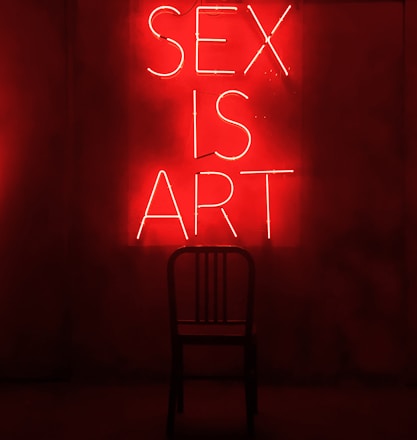 A dark room features a bright red neon sign displaying the words 'SEX IS ART'. In front of the sign, a black chair is silhouetted against the red glow.