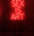 A dark room features a bright red neon sign displaying the words 'SEX IS ART'. In front of the sign, a black chair is silhouetted against the red glow.
