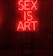 A dark room features a bright red neon sign displaying the words 'SEX IS ART'. In front of the sign, a black chair is silhouetted against the red glow.