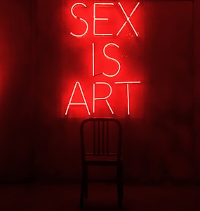 A dark room features a bright red neon sign displaying the words 'SEX IS ART'. In front of the sign, a black chair is silhouetted against the red glow.