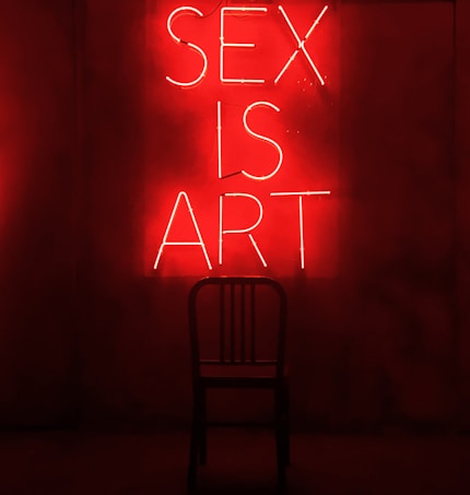 A dark room features a bright red neon sign displaying the words 'SEX IS ART'. In front of the sign, a black chair is silhouetted against the red glow.