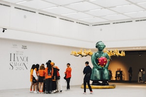 A group of people are gathered around an exhibition piece in an art gallery. The artwork features a large green statue with intricate details holding a red spherical object. Several individuals, dressed in bright orange outfits, are closely observing the art piece while another person stands nearby with their back turned. The gallery has minimalistic bright lighting and white walls with exhibition text displayed.