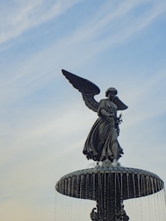 A bronze statue of an angel with outstretched wings stands atop a decorative fountain. Water gracefully cascades down from the fountain under a clear blue sky.