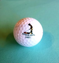 A golf ball with a textured surface placed on a light blue background. The ball features a logo of a person engaged in a golfing stance, accompanied by the text 'PINEHURST 1895'.