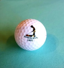 A golf ball with a textured surface placed on a light blue background. The ball features a logo of a person engaged in a golfing stance, accompanied by the text 'PINEHURST 1895'.